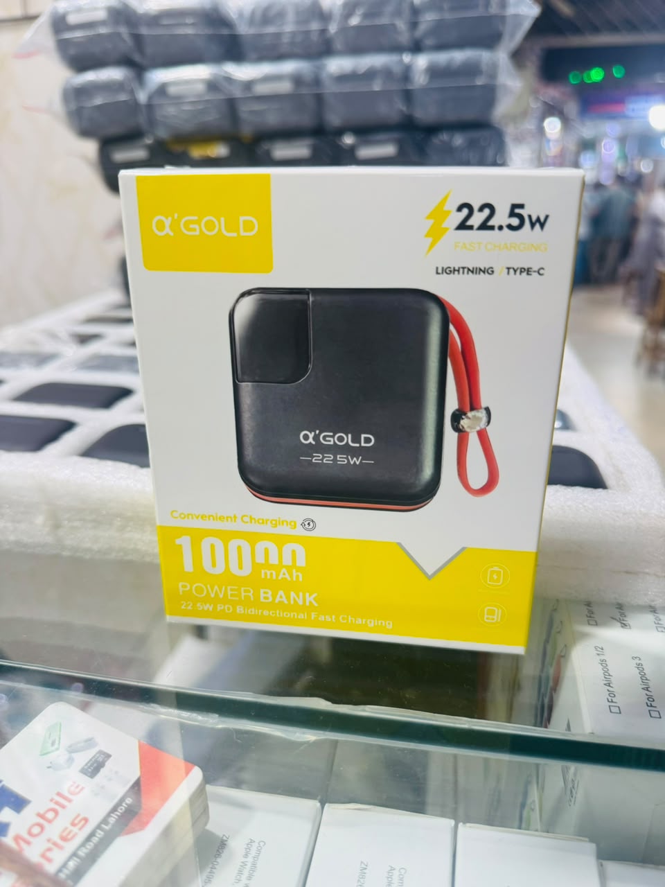 A boxed portable power bank on a glass display shelf. The packaging is white and yellow with a photo of a black square power bank and a red wrist strap, and text including “ΑΠΩL Gold,” “22.