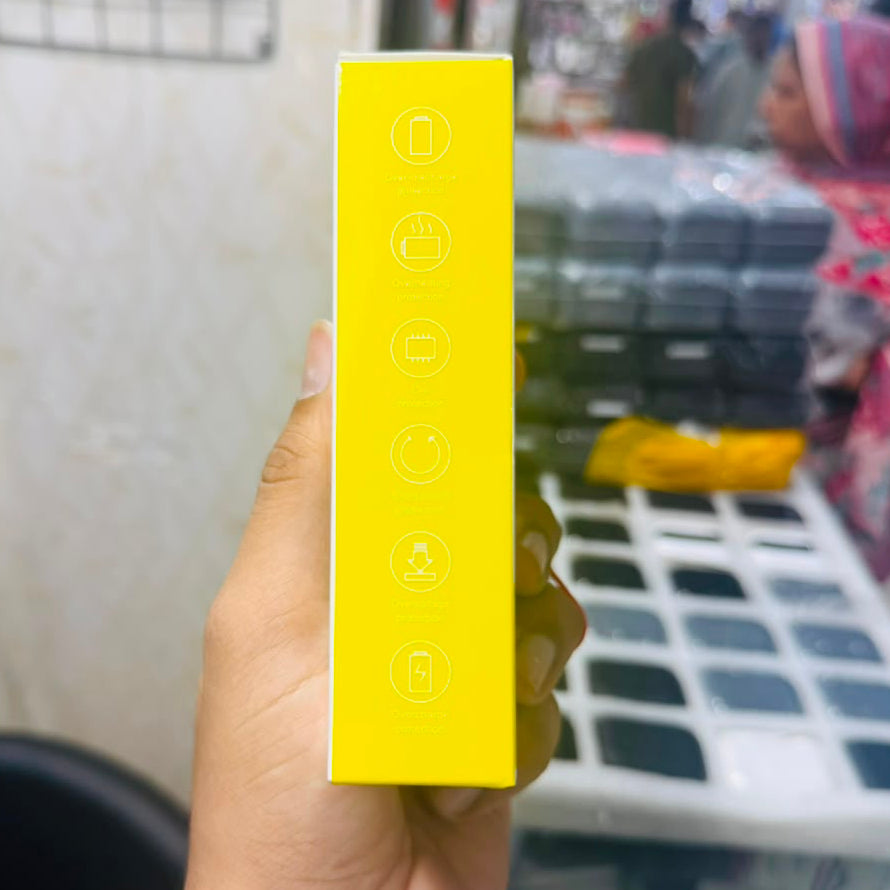 A hand holding a bright yellow rectangular product box with white icons printed on the side, photographed in a retail store environment.