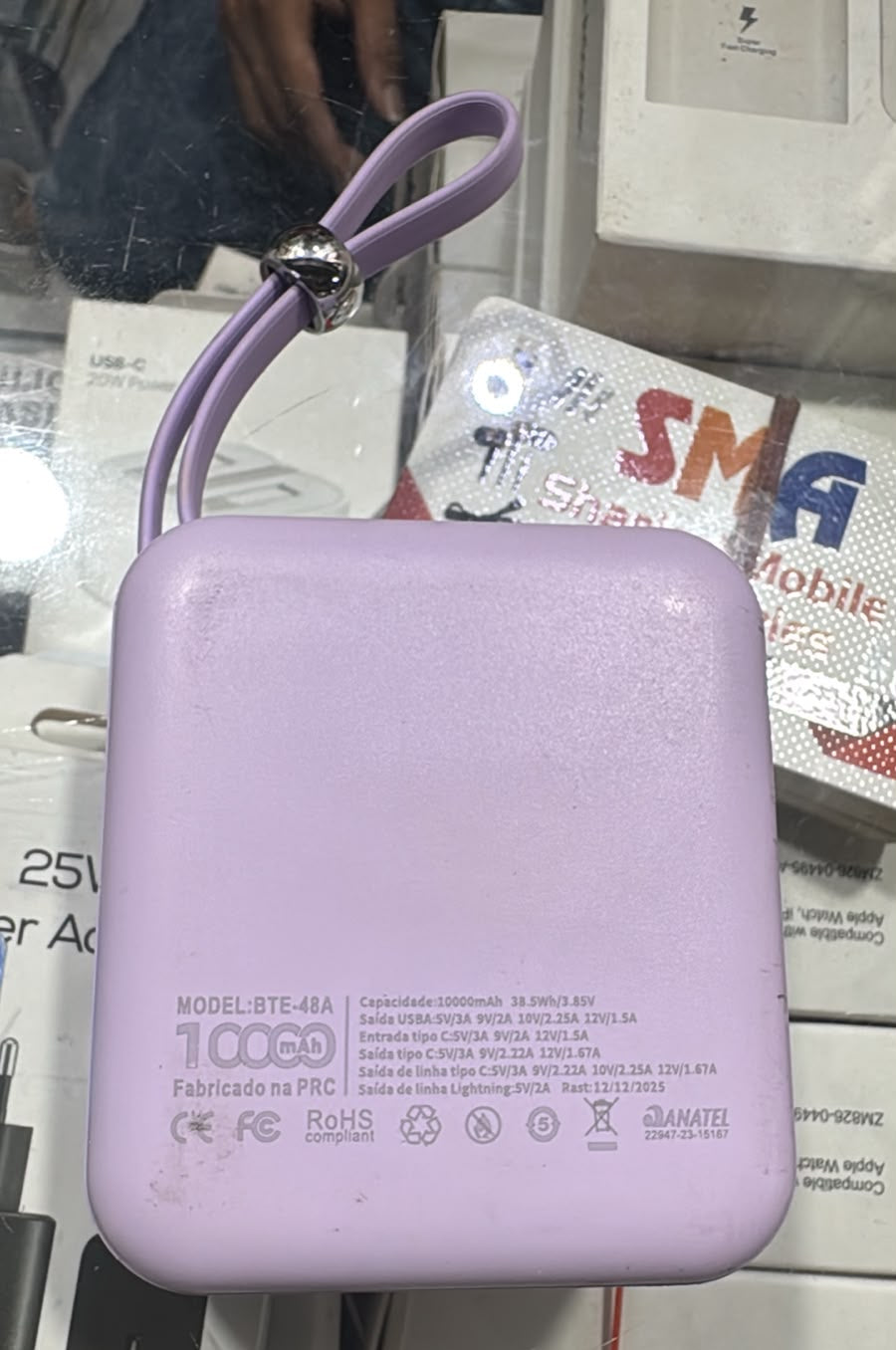 A light purple rectangular portable power bank with a matching wrist strap, displayed on top of its product packaging boxes.
