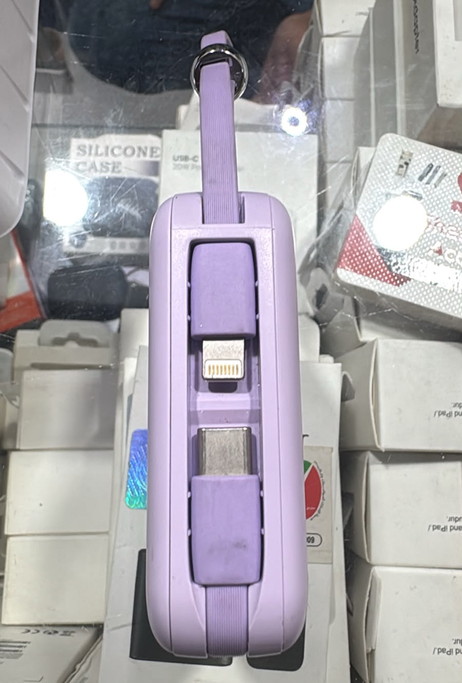 A lavender purple folding tablet case with built-in USB-C and Lightning ports, shown standing upright in a retail display surrounded by Samsung and other tech product boxes.