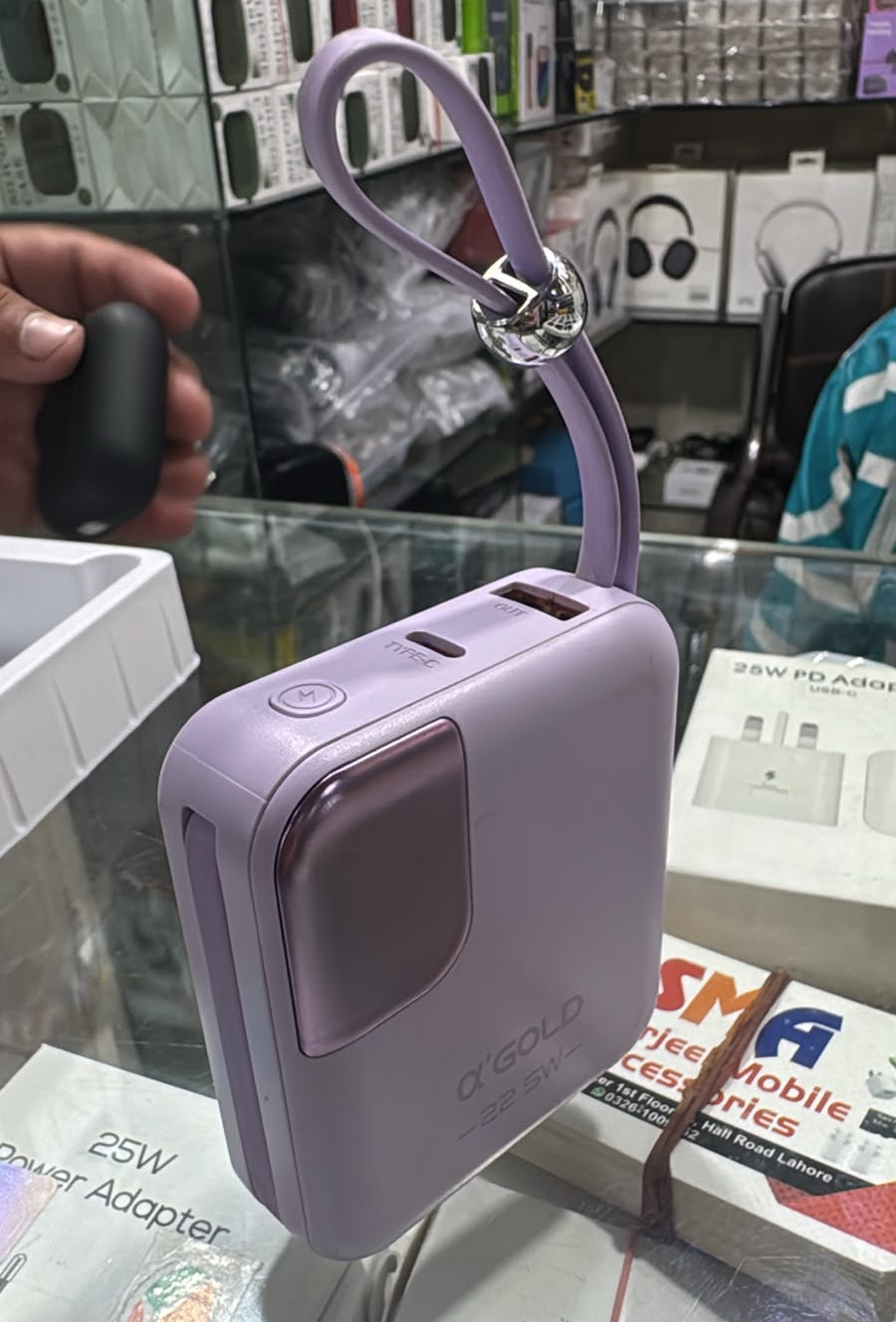 A lavender purple portable power bank with a matching wrist strap and silver clasp, displayed on a glass counter in a retail store. The brand 'samo' is visible on the front of the device.